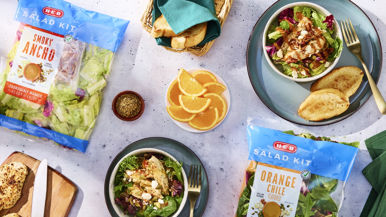 H‑E‑B Salad Kits in Smoky Ancho and Orange Chile flavors, prepared salads in bowls, garnished with cheese, served with toasted bread, sliced oranges, seasoning, a basket of bread, and sliced chicken on a cutting board.