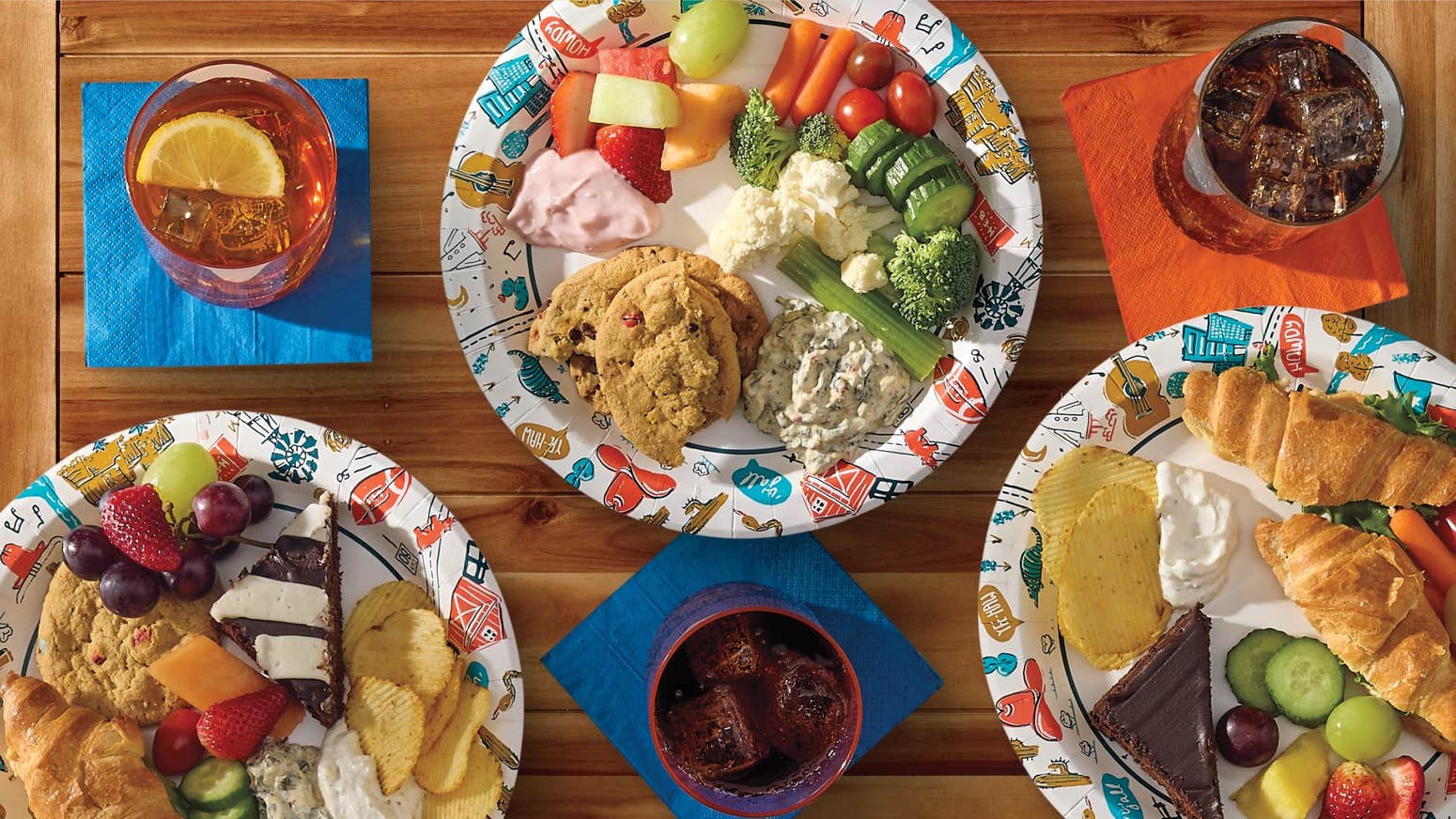 Four plates of assorted snacks—fruits, vegetables, croissants, and desserts—on a wooden table with three drinks.