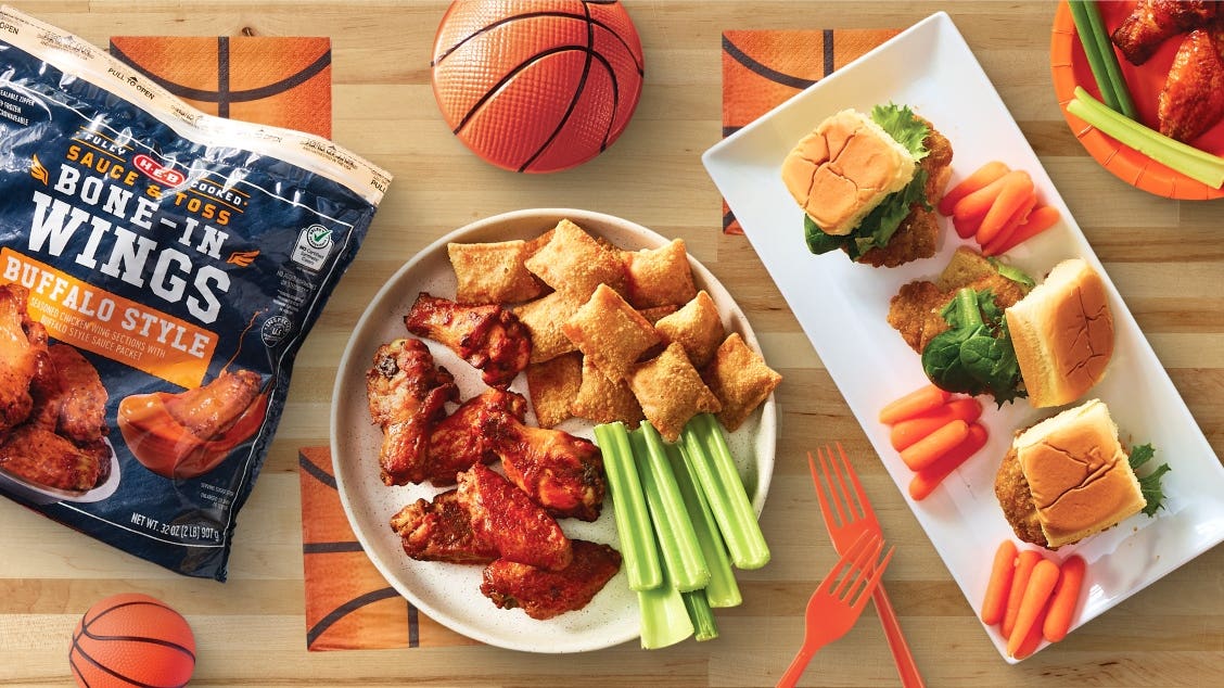 A table with basketball-themed decor displays buffalo wings, pizza rolls, sliders with lettuce, celery and carrot sticks, and mini basketballs.