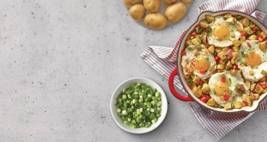 Breakfast skillet with sunny-side-up eggs, diced potatoes, and colorful bell peppers in a red pan, accompanied by a bowl of chopped green onions and fresh potatoes on a gray countertop.