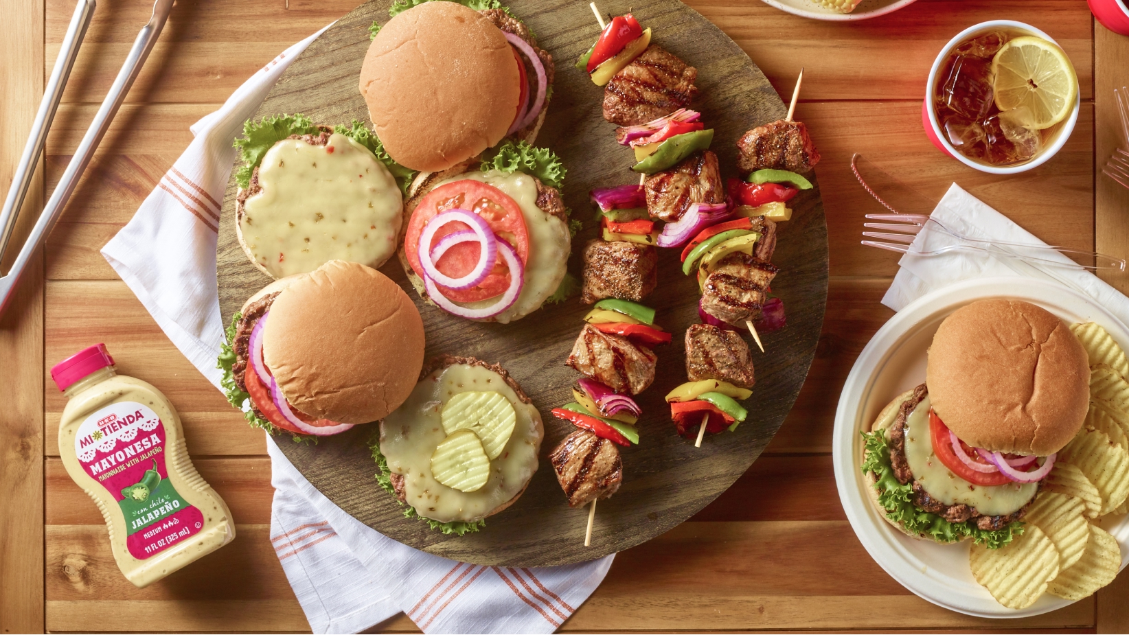 A delicious spread of grilled cheeseburgers with lettuce, tomato, and onion, accompanied by grilled vegetable and meat skewers, a bottle of jalapeño mayonnaise, a plate of chips, and a refreshing drink with lemon slices.
