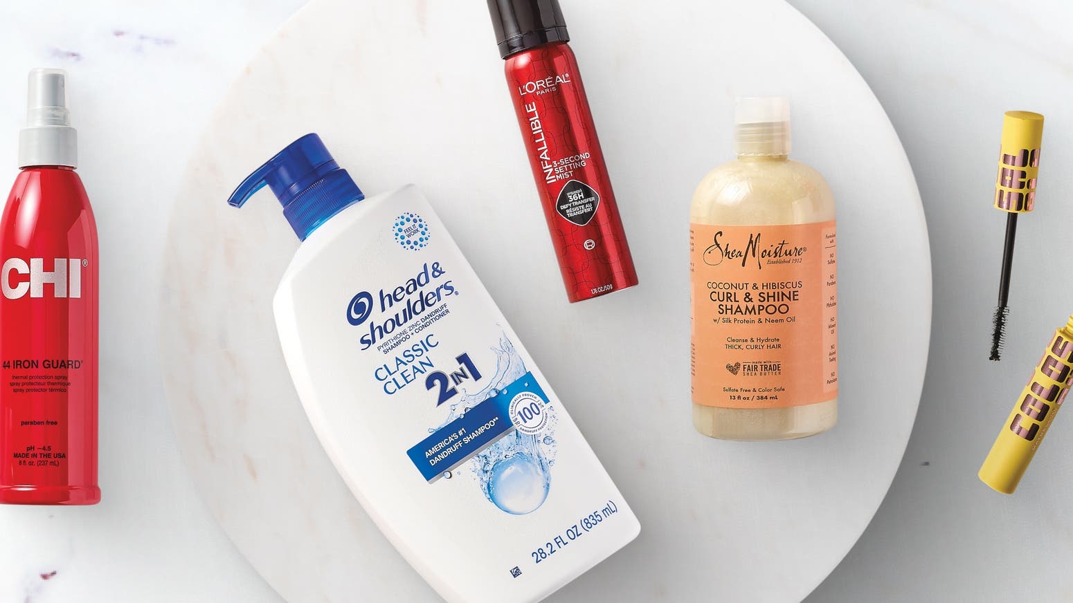 A selection of hair care products displayed on a marble surface, including CHI Iron Guard spray, Head & Shoulders Classic Clean 2-in-1 shampoo, L'Oréal Paris Infallible Hair Spray, Shea Moisture Curl & Shine Shampoo, and a Colossal Volume Mascara.