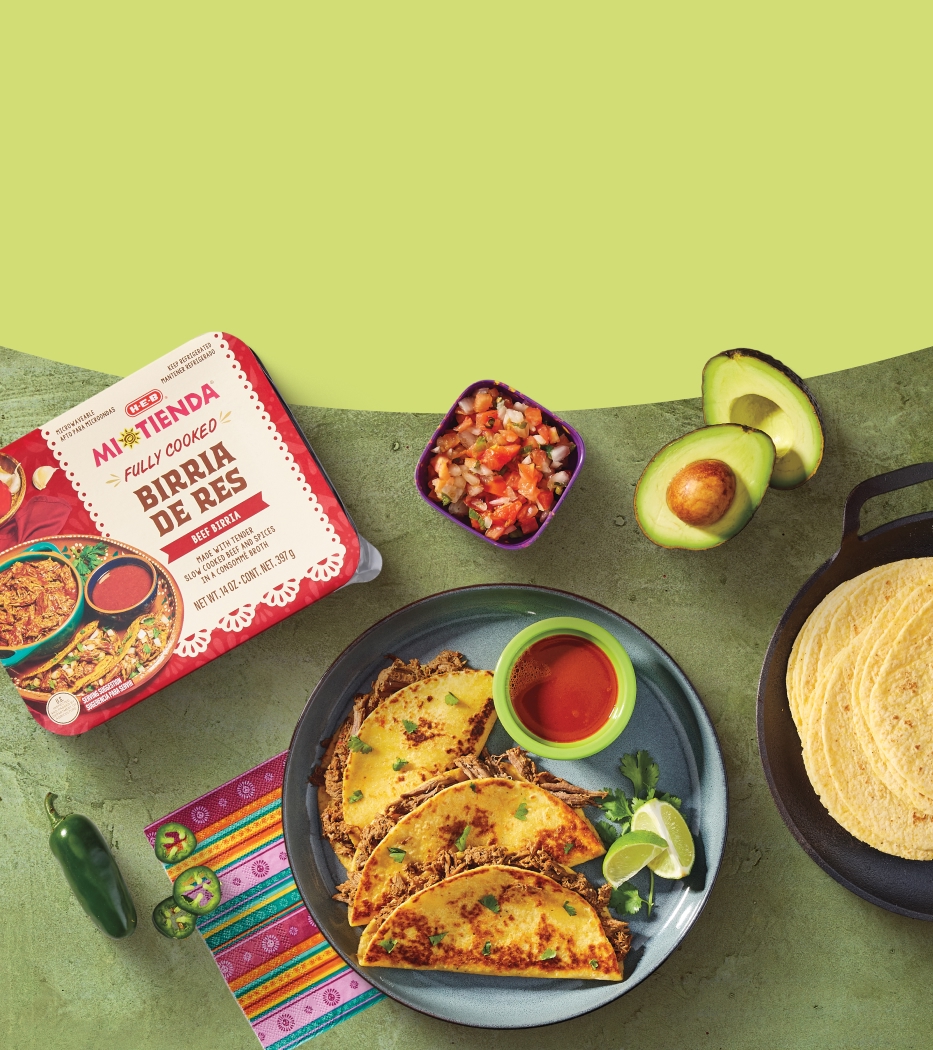 Mexican food spread featuring beef birria tacos, tortillas, avocado, salsa, and jalapeño, with a Mi Tienda ready-to-cook beef birria package on a green background.