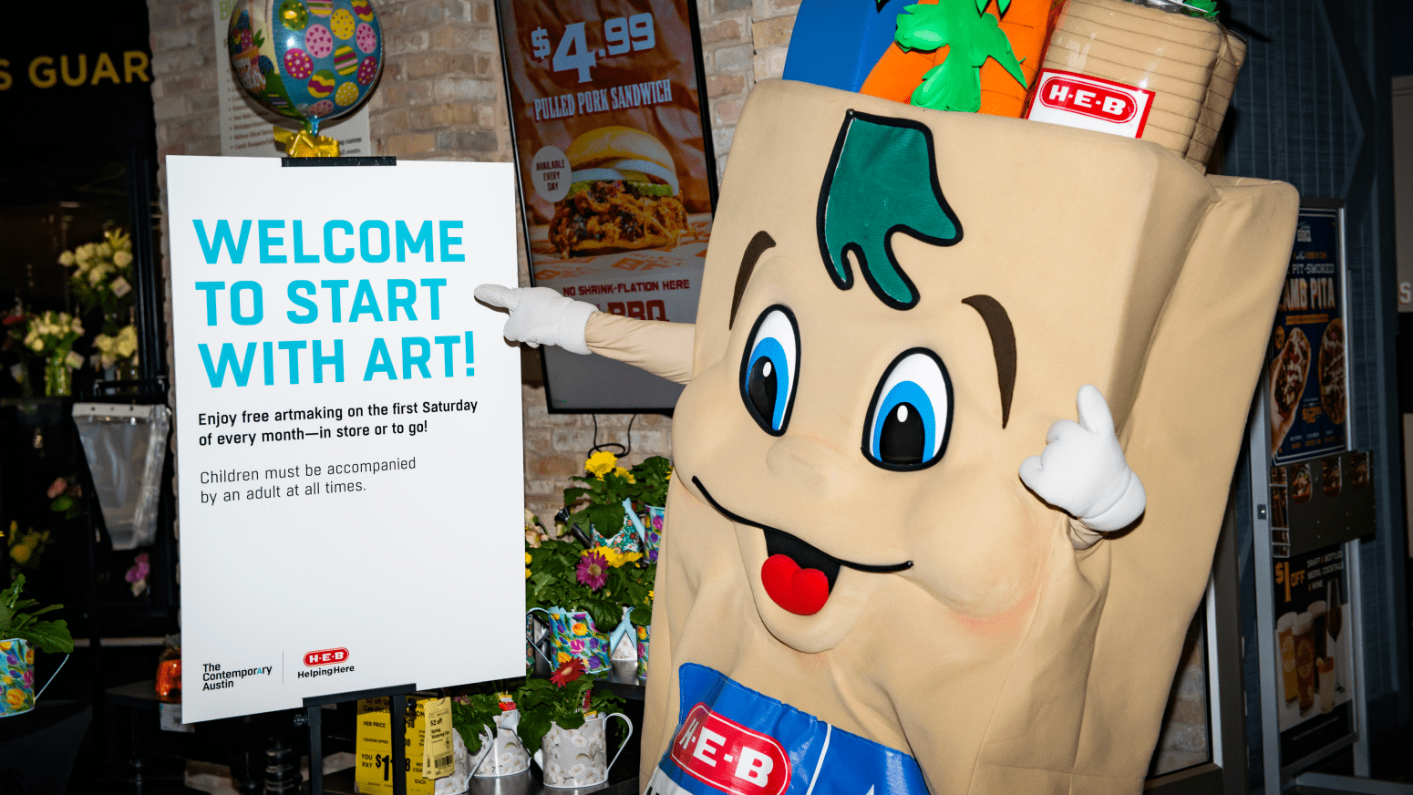 H‑E‑Buddy mascot dressed as a grocery bag with a smiling face and H‑E‑B logo stands next to a sign promoting the 'Start With Art!' event, inviting families to enjoy free artmaking on the first Saturday of every month.