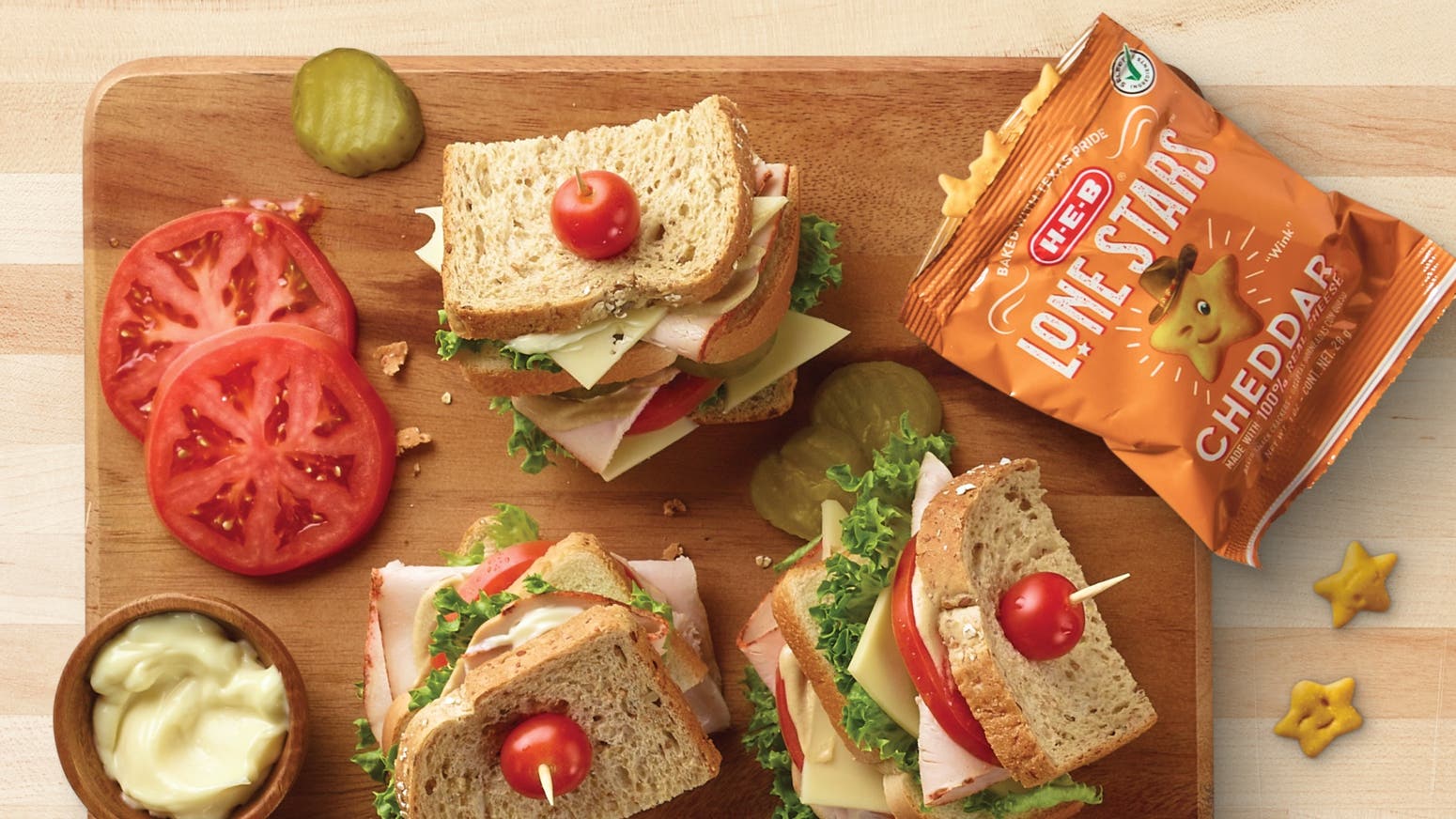H‑E‑B Mini Peanut Butter Sandwich Crackers, H‑E‑B Lone Stars cheddar crackers, and two sandwiches with tomato and pickle on a wooden cutting board.