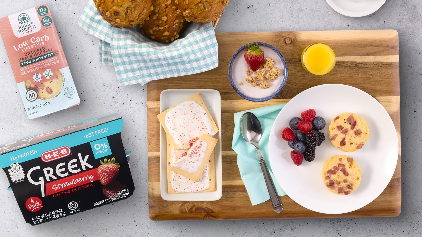 Breakfast spread featuring H‑E‑B Greek Strawberry Yogurt, Higher Harvest Low-Carb Egg White Bites, assorted berries, muffins, and a small glass of orange juice on a wooden board.