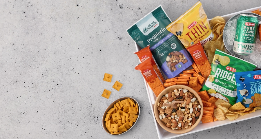 A variety of H‑E‑B snack products including chips, crackers, trail mix, and canned drinks displayed on a light gray surface.