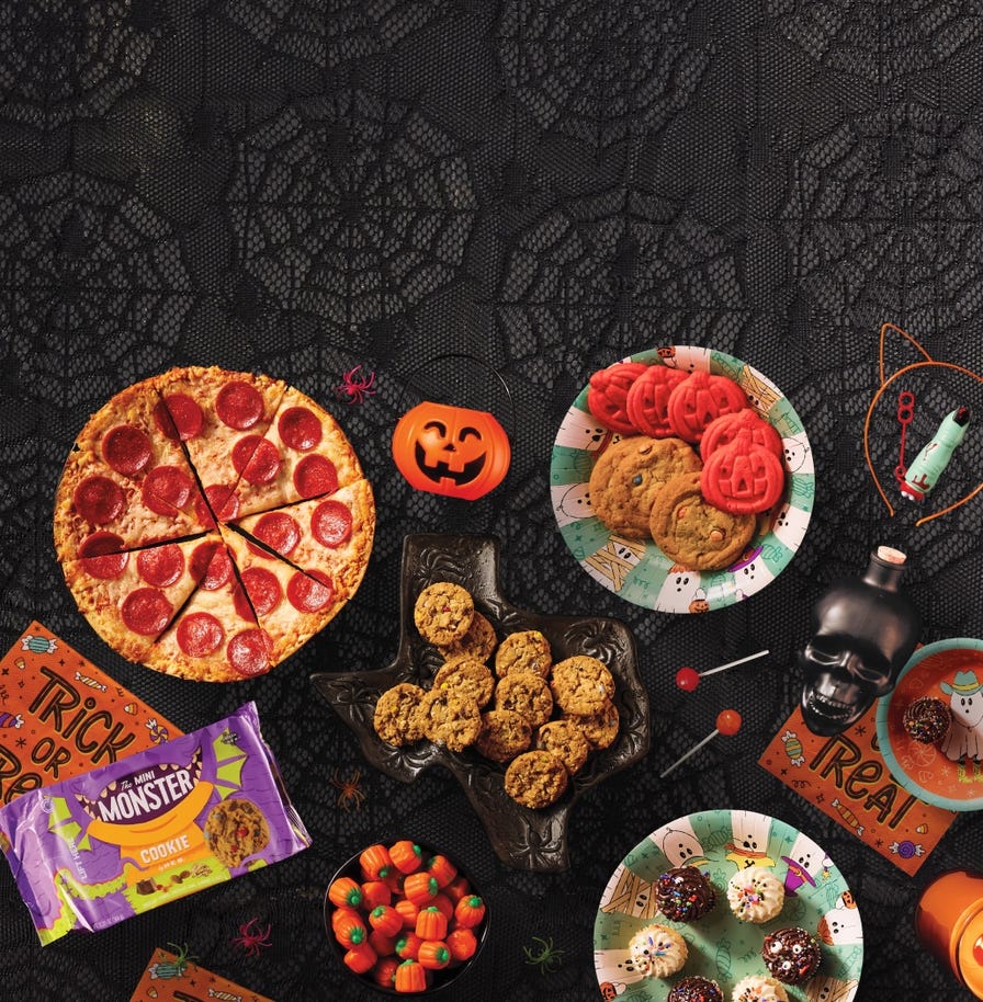Halloween party food spread featuring a pepperoni pizza, assorted cookies, cupcakes, candy, and festive decorations on a spider web tablecloth.