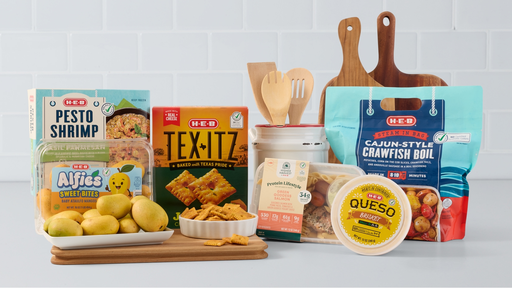 H‑E‑B branded food products displayed on a kitchen counter, including Pesto Shrimp, Tex-Itz crackers, Cajun-Style Crawfish Boil, Alfies Sweet Bites mangoes, Green Goddess Salmon, and Queso, along with wooden cutting boards and kitchen utensils.