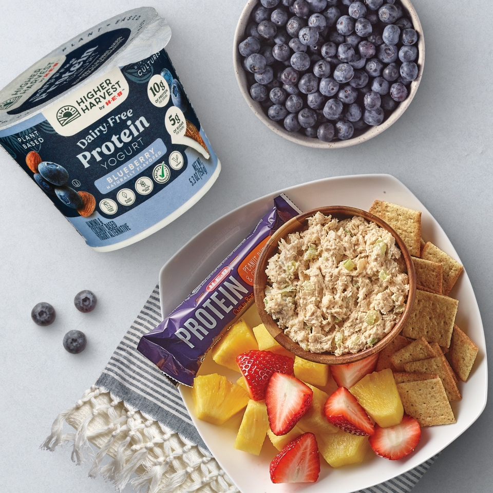 H‑E‑B Higher Harvest Dairy-Free Blueberry Protein Yogurt, fresh blueberries, chicken salad with crackers, pineapple, strawberries, and a protein bar on a table.