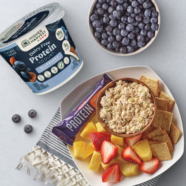 H‑E‑B Higher Harvest Dairy-Free Blueberry Protein Yogurt, fresh blueberries, chicken salad with crackers, pineapple, strawberries, and a protein bar on a table.