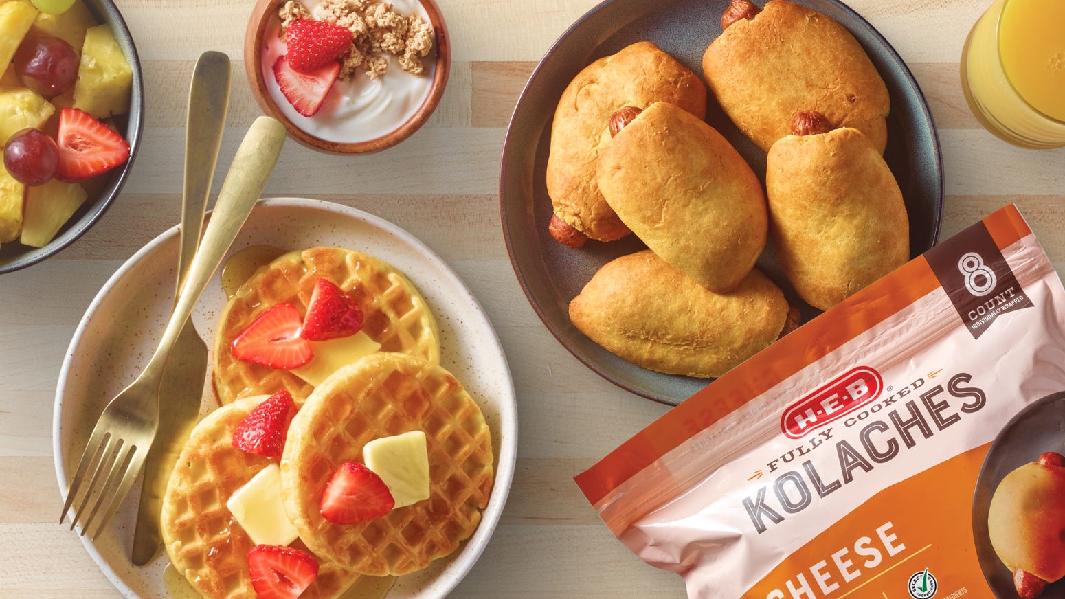 H‑E‑B Fully Cooked Kolaches next to plates of waffles with fruit and butter, and a bowl of fruit and yogurt, for breakfast.