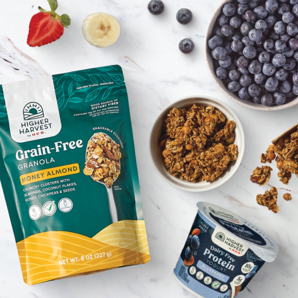 Higher Harvest by H‑E‑B Grain-Free Honey Almond Granola bag and bowl, Dairy-Free Blueberry Protein Yogurt cup, fresh blueberries, strawberries, and bananas on a marble surface.
