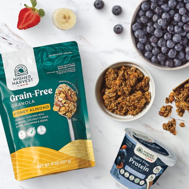 Higher Harvest by H‑E‑B Grain-Free Honey Almond Granola bag and bowl, Dairy-Free Blueberry Protein Yogurt cup, fresh blueberries, strawberries, and bananas on a marble surface.