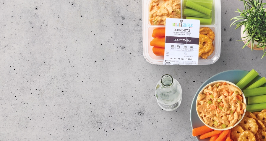 Buffalo-style chicken salad, celery sticks, carrots, pretzels, and a bottle of water on a concrete surface. Ready-to-eat packaged meal from Meal Simple.