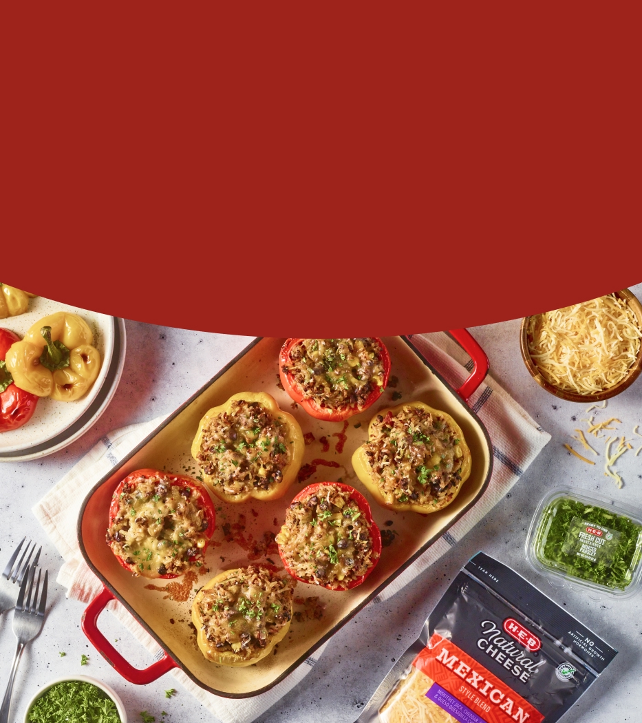 Baked stuffed bell peppers with melted cheese in a red baking dish, surrounded by ingredients including shredded cheese, pesto, and fresh herbs on a white countertop.