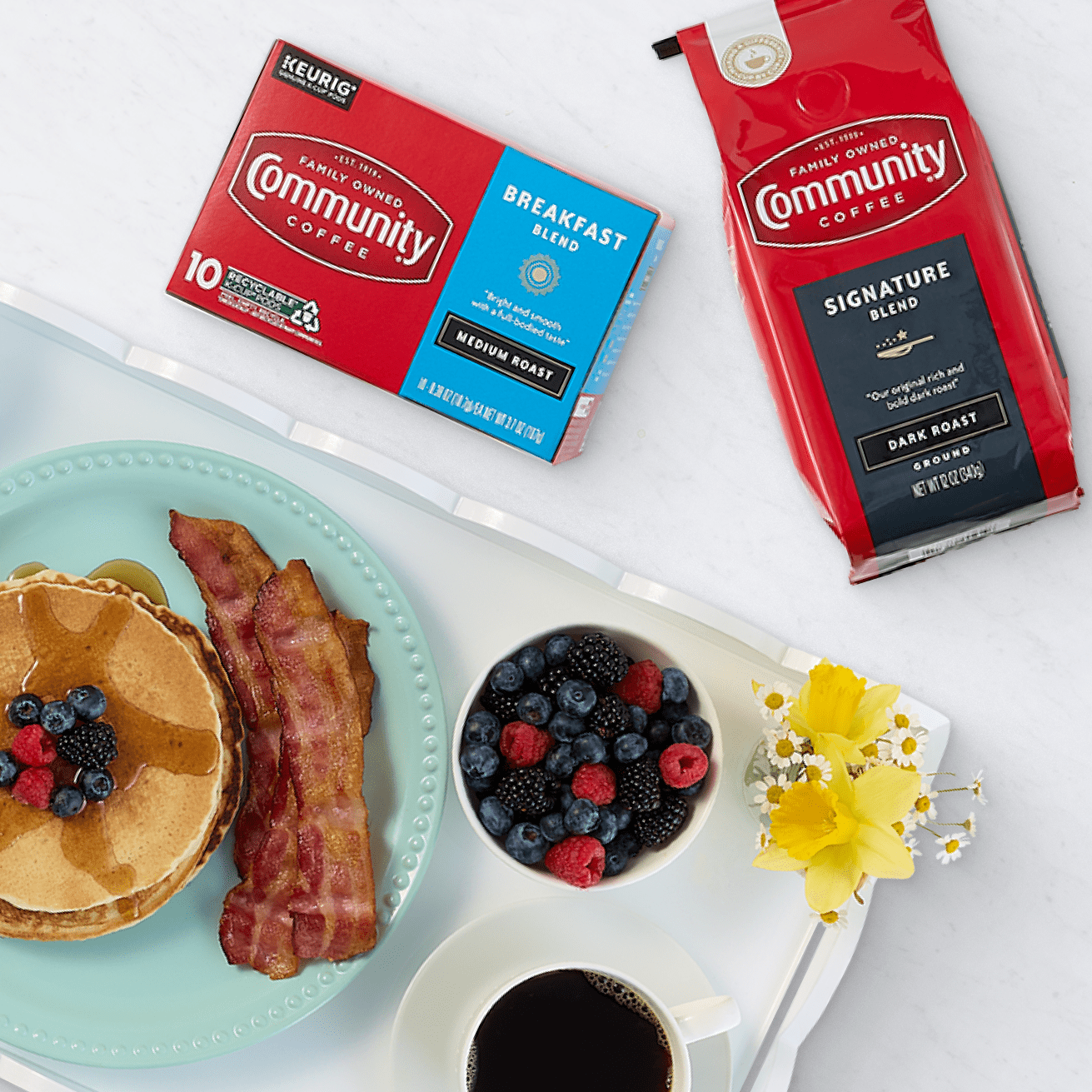 Pancakes topped with fresh berries, crispy bacon, a bowl of mixed berries, and a cup of coffee on a tray. Community Coffee's Breakfast Blend and Signature Blend packages are displayed in the background. 