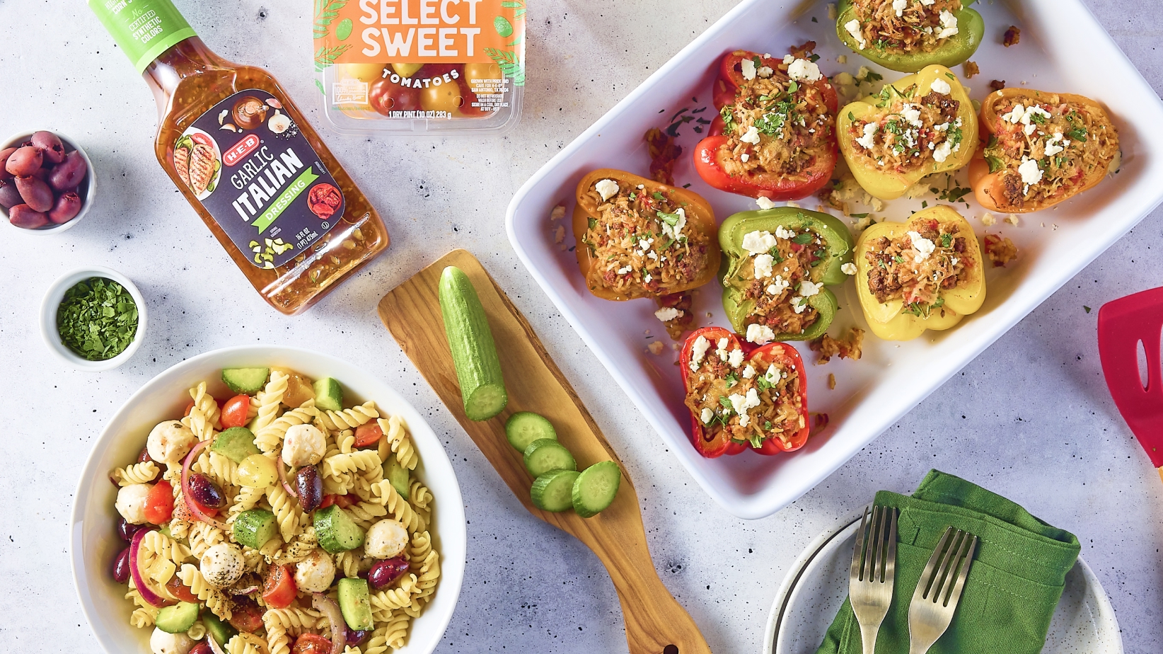 A bowl of pasta salad with vegetables and mozzarella, a tray of stuffed bell peppers topped with cheese, a bottle of garlic Italian dressing, and fresh ingredients like cucumbers and herbs.