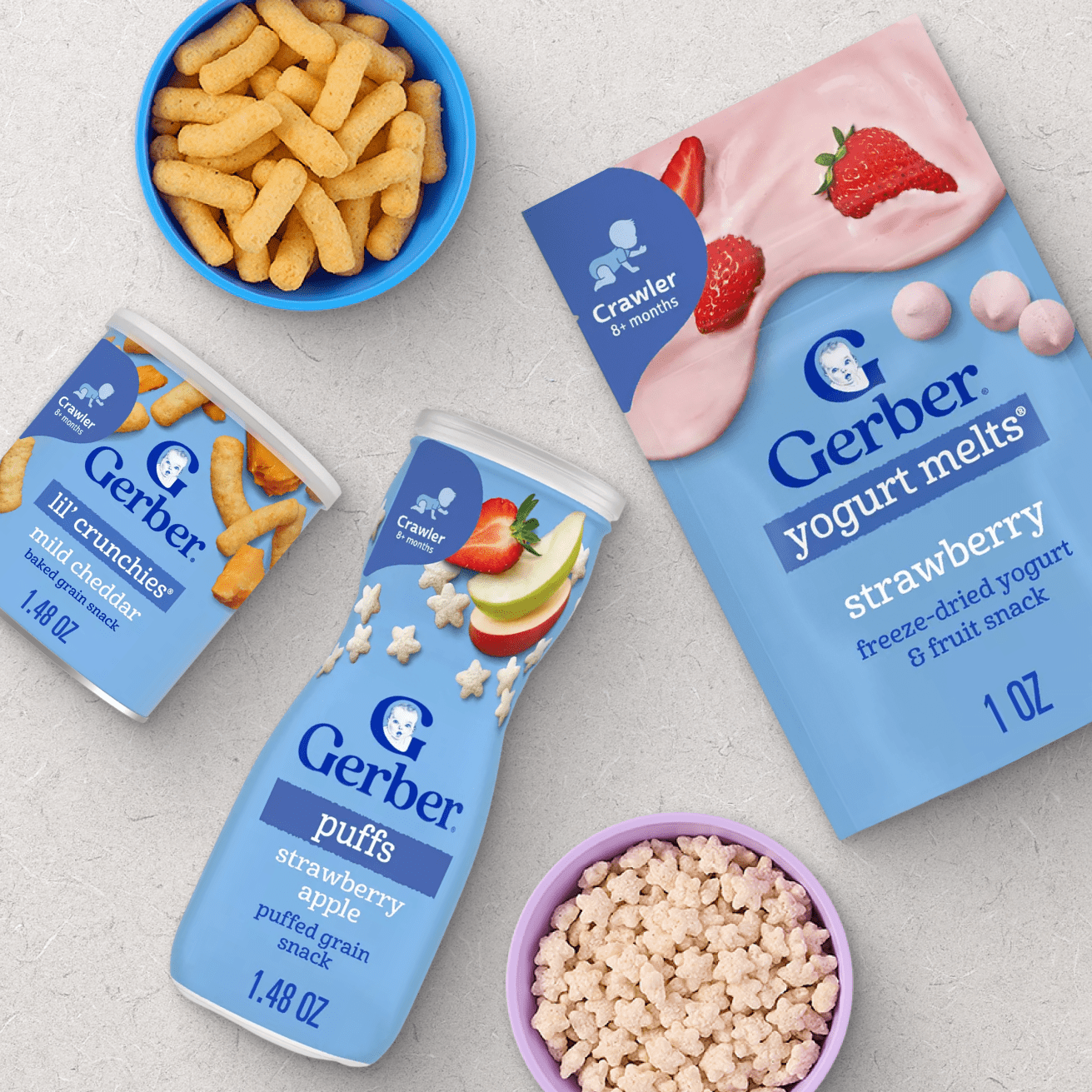 Gerber baby snacks assortment including Lil' Crunchies mild cheddar, Puffs strawberry apple, and Yogurt Melts strawberry displayed in blue and purple bowls on a light background.