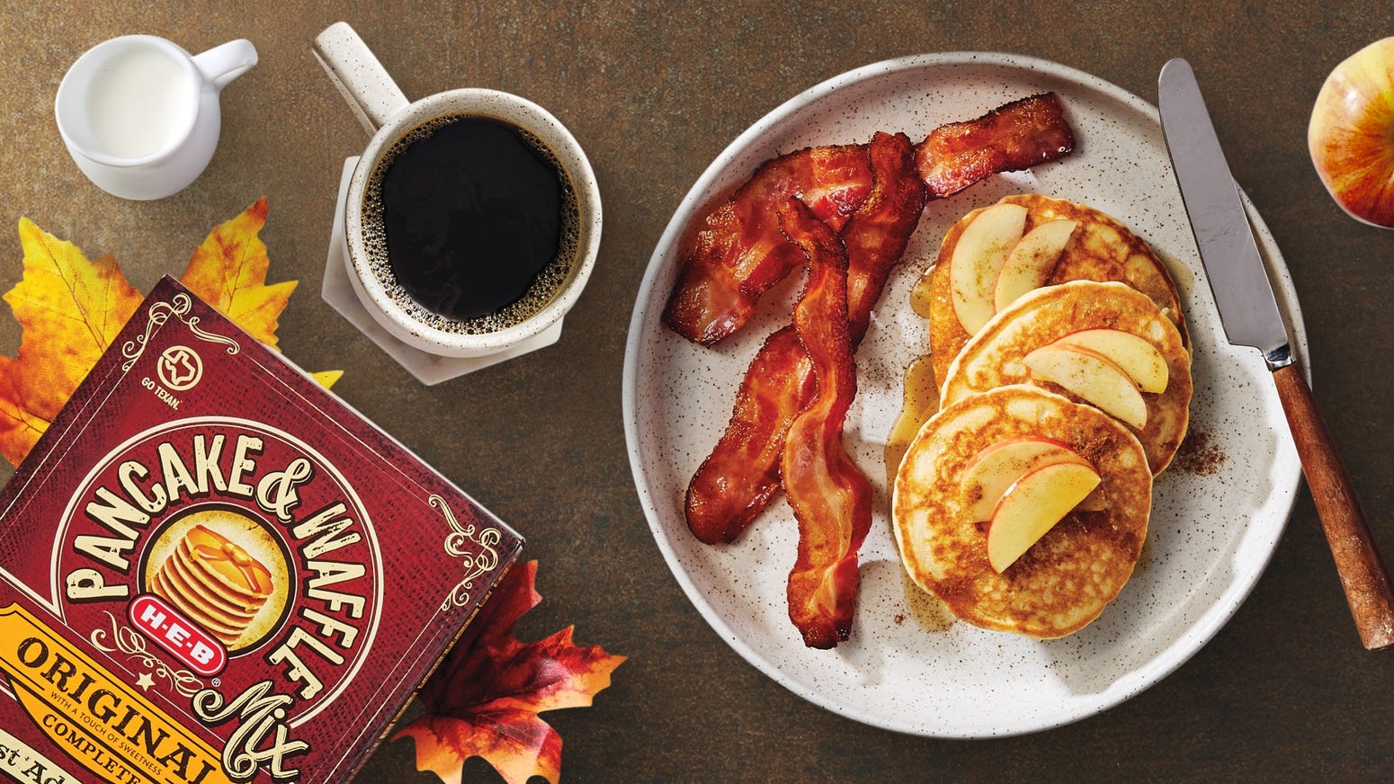 H‑E‑B Pancake & Waffle Mix, crispy bacon, apple-topped pancakes, black coffee, and milk, set on a rustic table with autumn leaves.