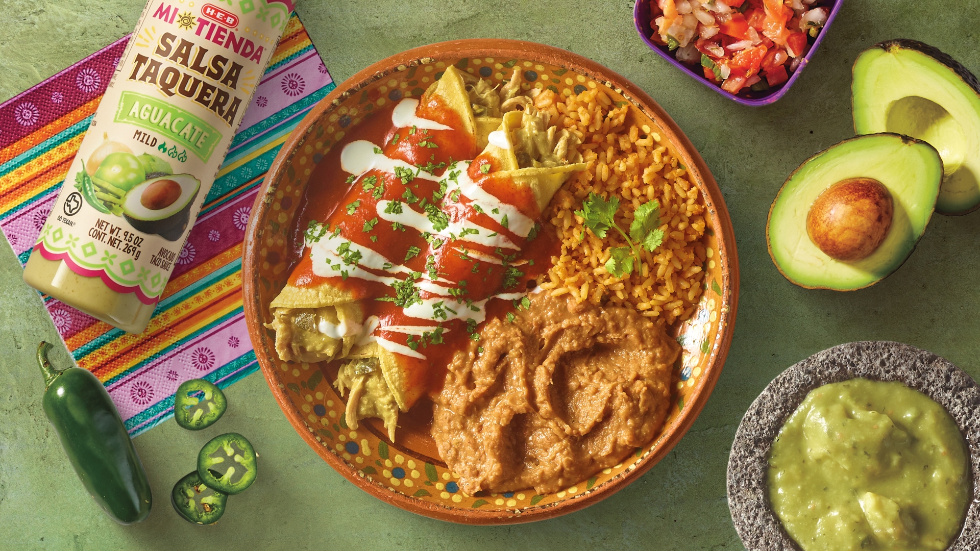 Mexican enchiladas topped with red sauce and sour cream, served with rice and refried beans, accompanied by H‑E‑B Mi Tienda Salsa Taquera Aguacate and a bowl of guacamole on a colorful tablecloth.