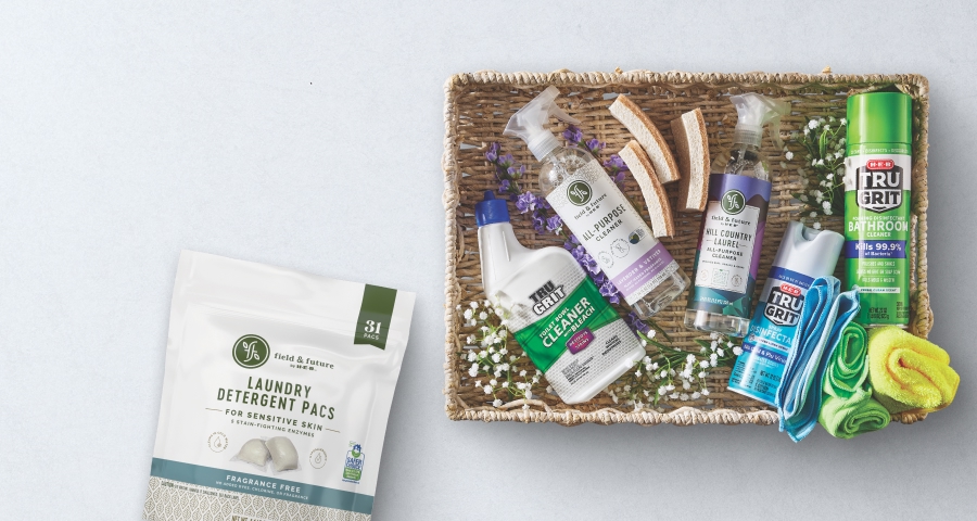 A variety of eco-friendly cleaning products, including Field & Future laundry detergent pacs, Tru Grit bathroom cleaner, and other household cleaners, displayed in a wicker basket with sponges and flowers on a light background.