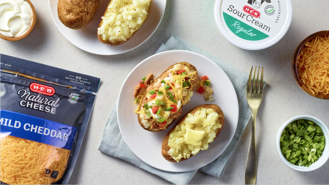 a spread of baked potatoes and various toppings, including H‑E‑B Sour Cream, H‑E‑B Natural Mild Cheddar shredded cheese, green onions, and butter.