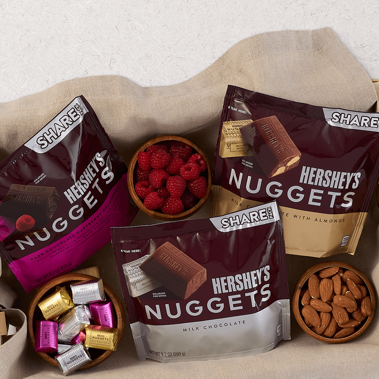 Assorted Hershey's Nuggets chocolate bags with bowls of raspberries, almonds, and wrapped chocolates on a beige cloth background.
