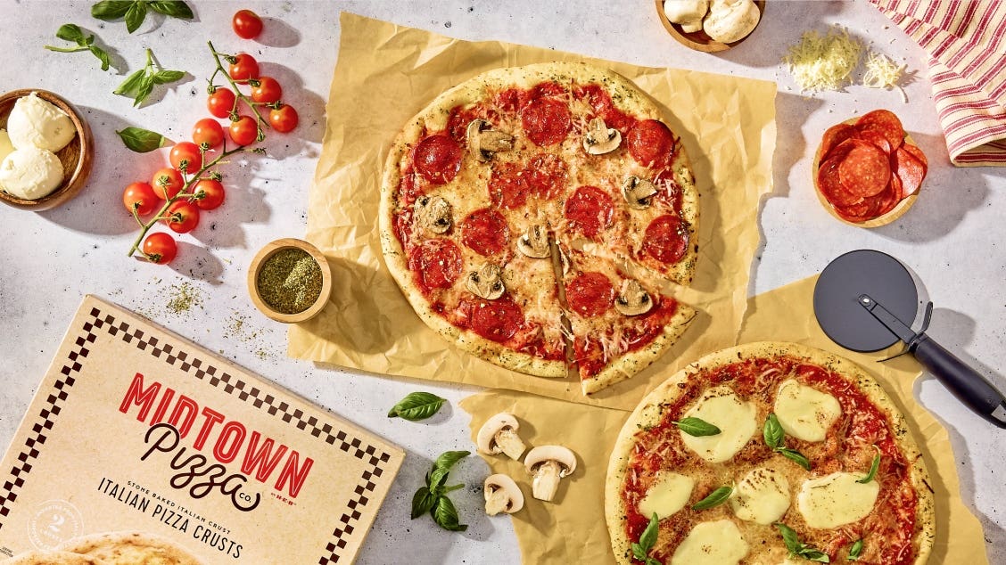 Two pizzas on parchment paper: one topped with pepperoni and mushrooms, another with mozzarella slices. Nearby are fresh tomatoes, mushroom slices, basil, grated cheese, Midtown Pizza box of crusts, and a pizza cutter.