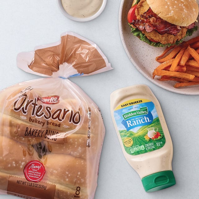 A meal featuring a crispy chicken sandwich with bacon, lettuce, and tomato on a plate with sweet potato fries. Nearby are Sara Lee Artesano Bakery Buns and Hidden Valley Ranch dressing.